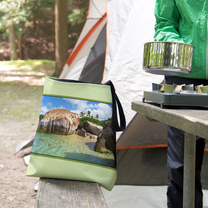 A tote bag featuring the calm turquoise water design sits on the bench of a campsite picnic table as the camper cooks.
