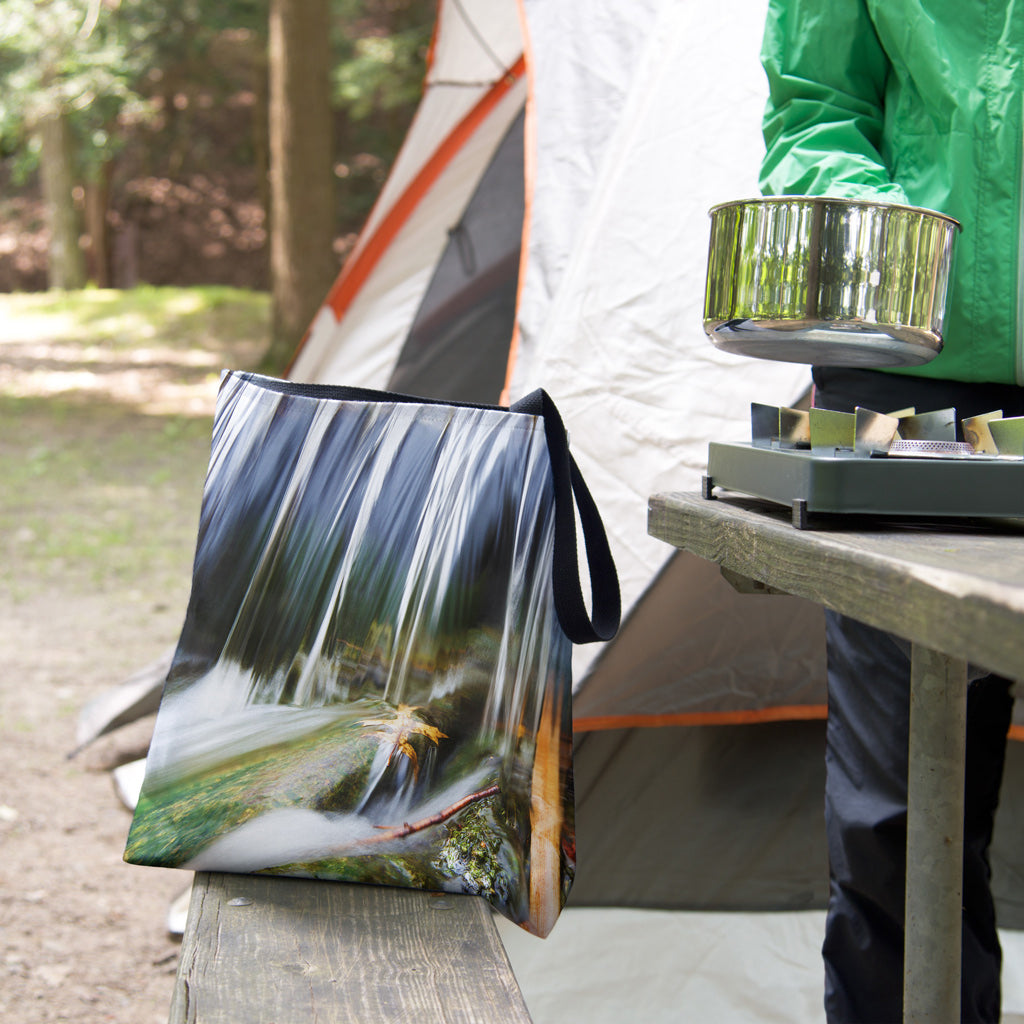 A tote bag featuring the creek waterfall design sits on the bench of a campsite picnic table as the camper cooks.