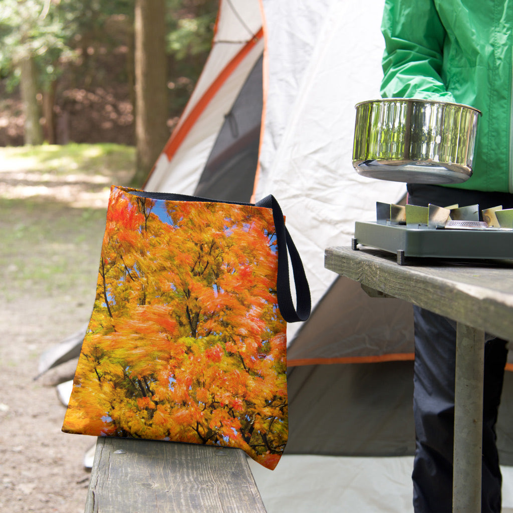 A tote bag featuring the fall colored trees design sits on the bench of a campsite picnic table as the camper cooks.