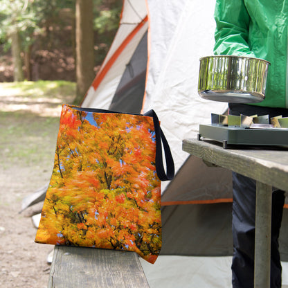 A tote bag featuring the fall colored trees design sits on the bench of a campsite picnic table as the camper cooks.