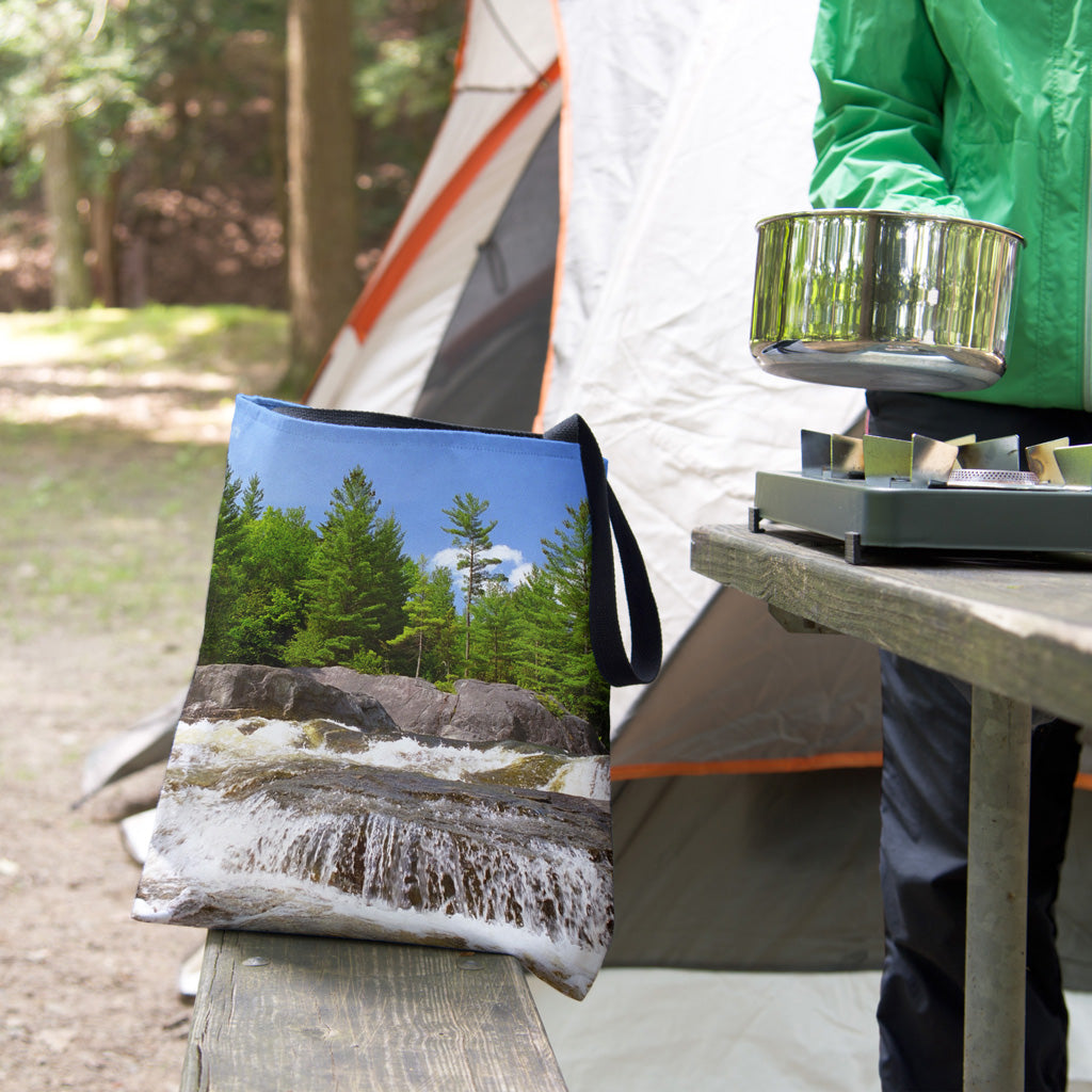 A tote bag featuring the forest waterfall design sits on the bench of a campsite picnic table as the camper cooks.