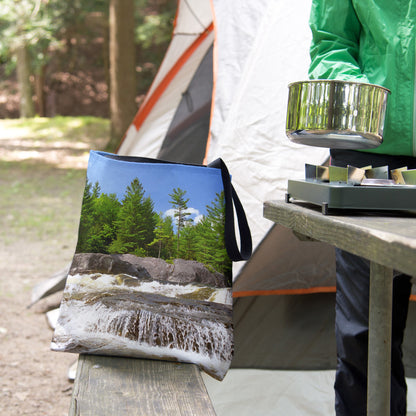 A tote bag featuring the forest waterfall design sits on the bench of a campsite picnic table as the camper cooks.
