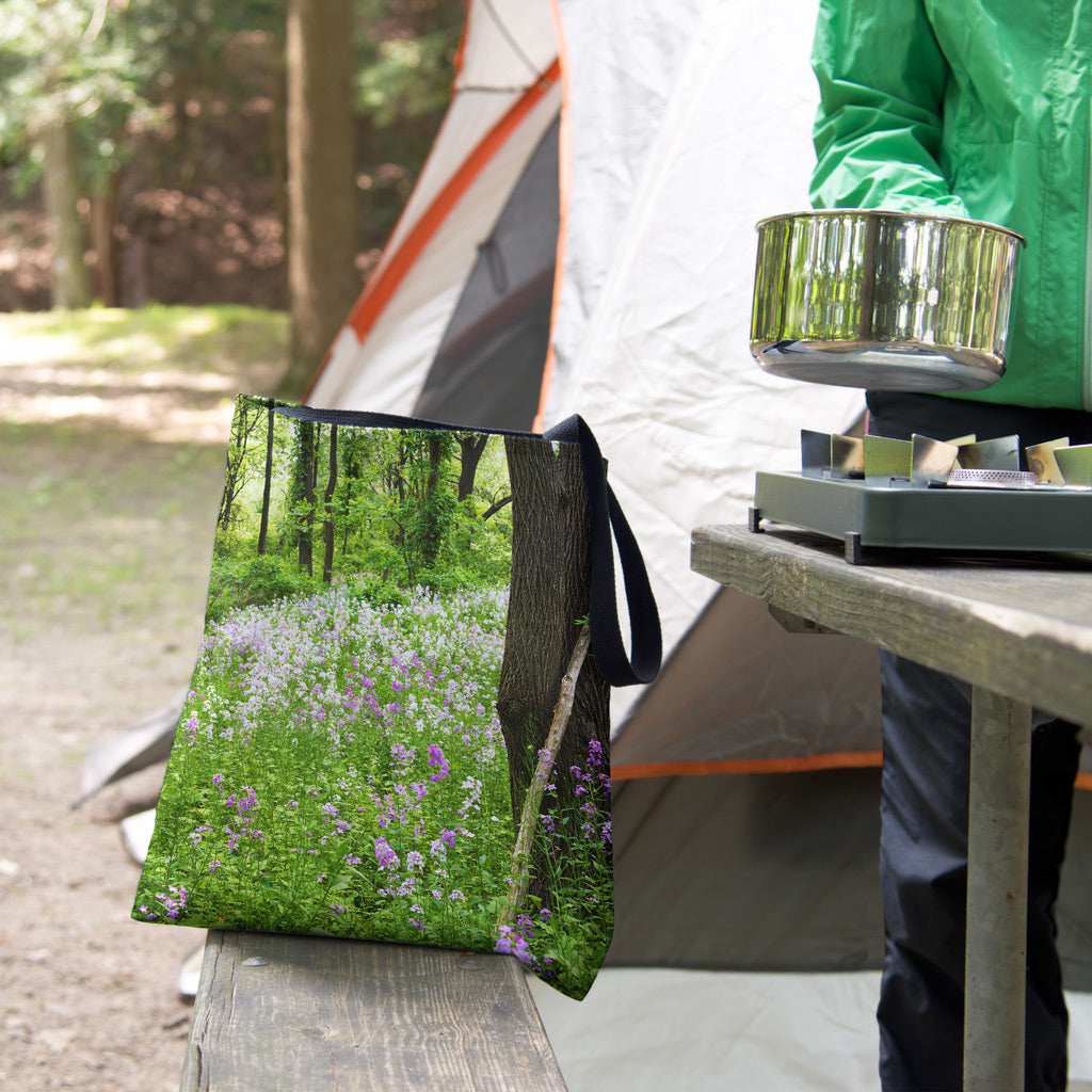A tote bag featuring the forest wildflowers design sits on the bench of a campsite picnic table as the camper cooks.