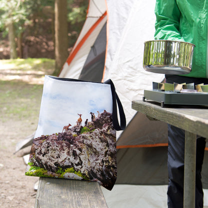 A tote bag featuring the curious goats design sits on the bench of a campsite picnic table as the camper cooks.