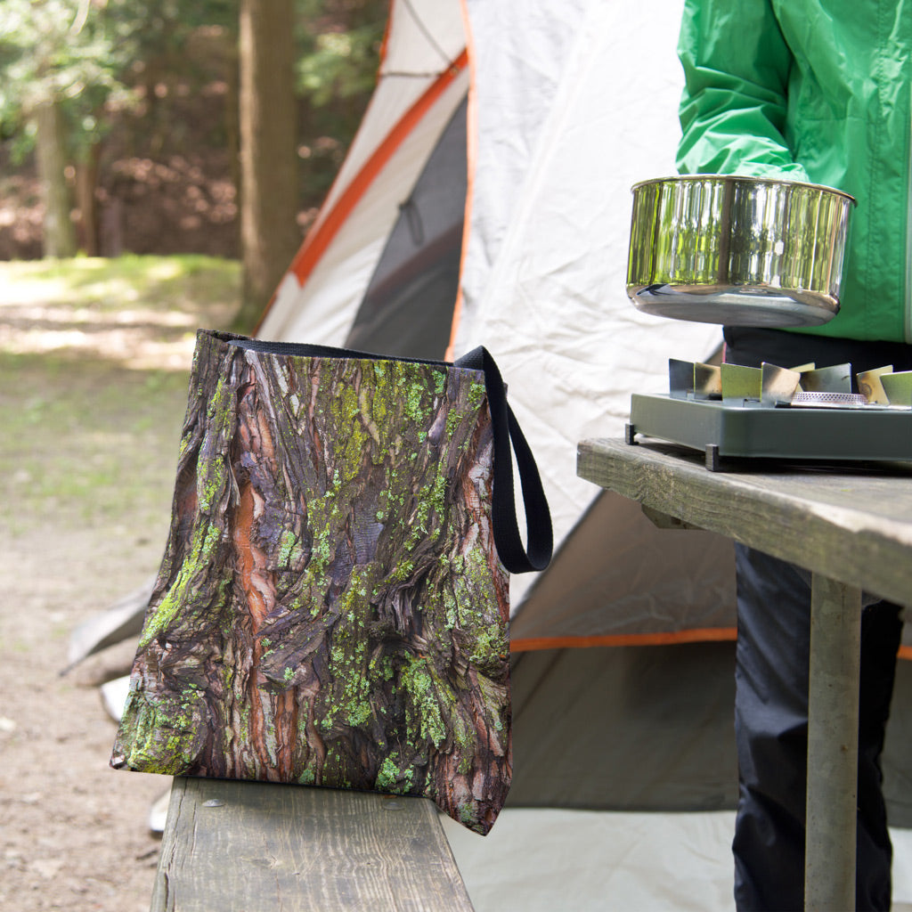A tote bag featuring the green mossy bark design sits on the bench of a campsite picnic table as the camper cooks.