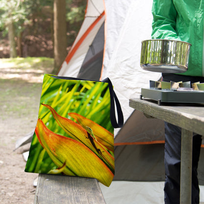 A tote bag featuring the sunlit gecko design sits on the bench of a campsite picnic table as the camper cooks.