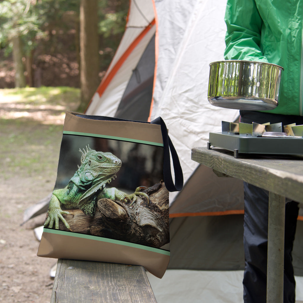 A tote bag featuring the green iguana design sits on the bench of a campsite picnic table as the camper cooks.