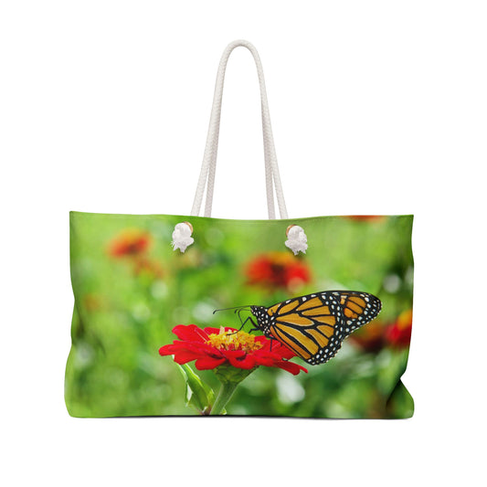 Front view of a beach bag featuring vibrant photo art close up of a monarch butterfly perched on a large red flower.
