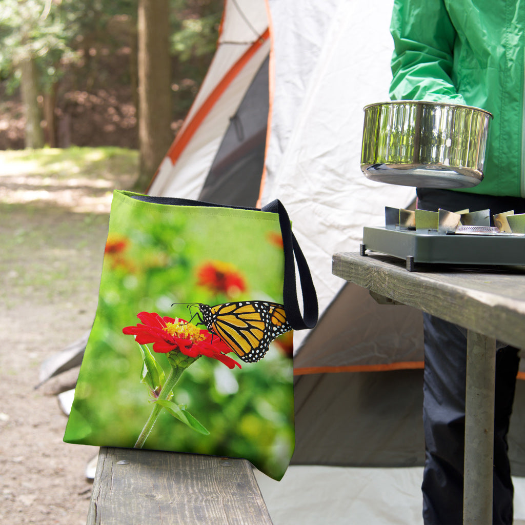 A tote bag featuring the butterfly on flower design sits on the bench of a campsite picnic table as the camper cooks.
