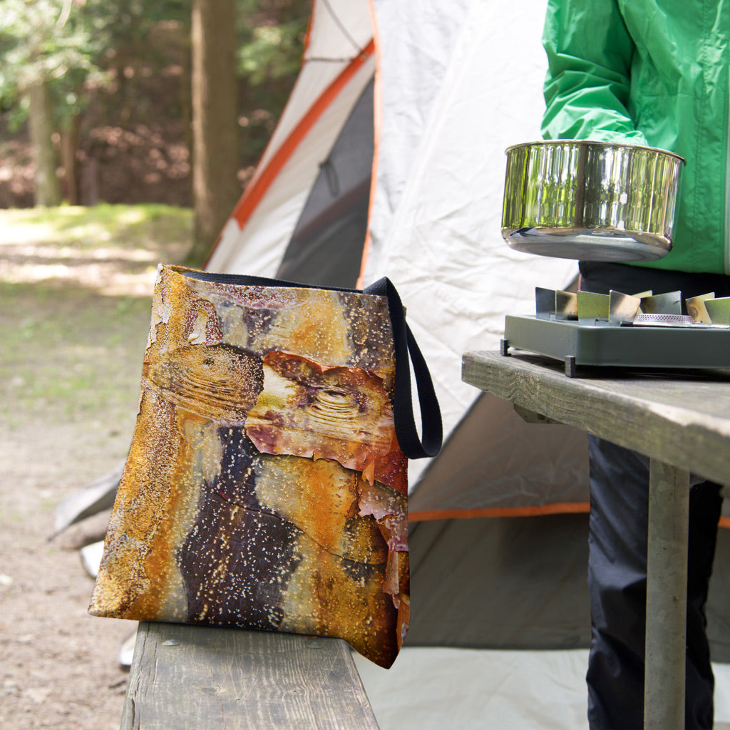 A tote bag featuring the orange bark design sits on the bench of a campsite picnic table as the camper cooks.