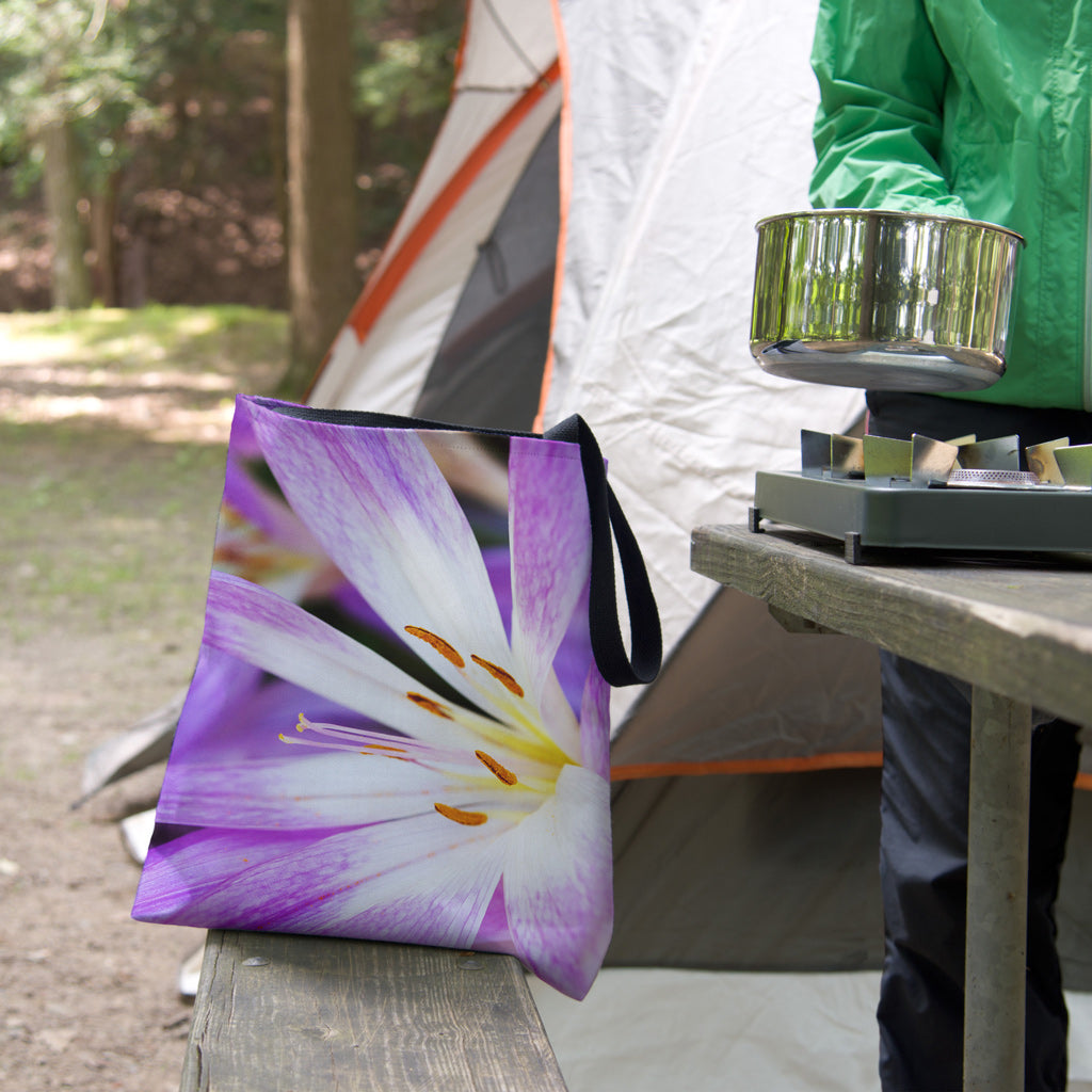 A tote bag featuring the purple flower, orange stamen design sits on the bench of a campsite picnic table as the camper cooks.