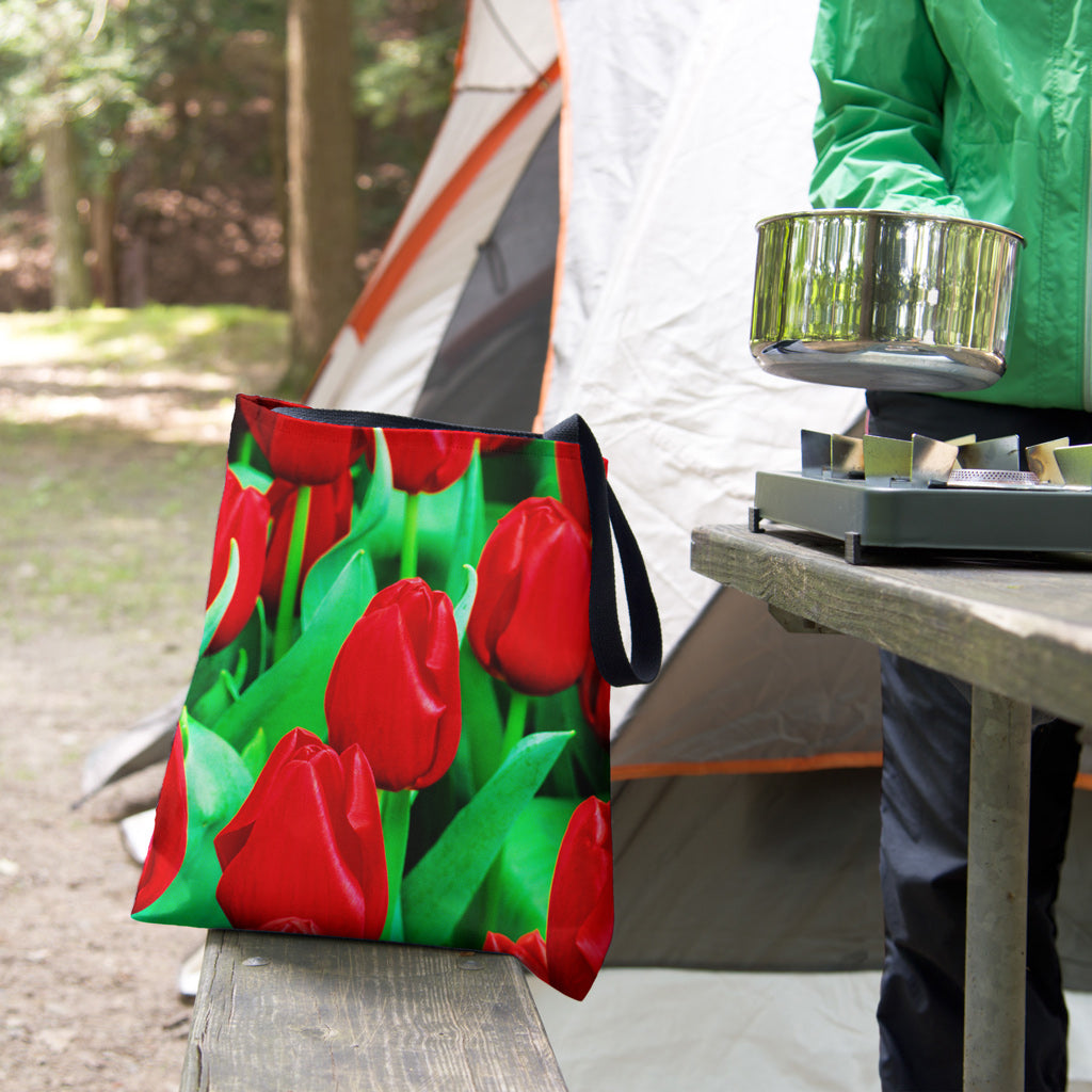 A tote bag featuring the red tulips design sits on the bench of a campsite picnic table as the camper cooks.