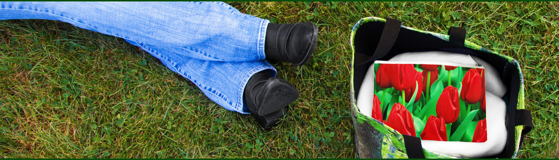 Person in blue jeans and black boots lying in the grass next to a bag with red tulip design.