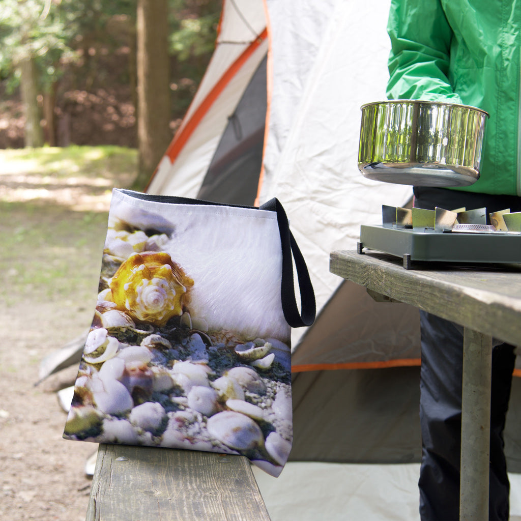 A tote bag featuring the shell in surf design sits on the bench of a campsite picnic table as the camper cooks.