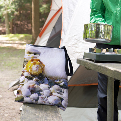 A tote bag featuring the shell in surf design sits on the bench of a campsite picnic table as the camper cooks.