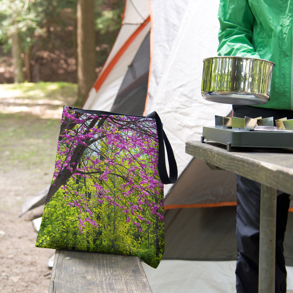 A tote bag featuring the tree blossoms design sits on the bench of a campsite picnic table as the camper cooks.