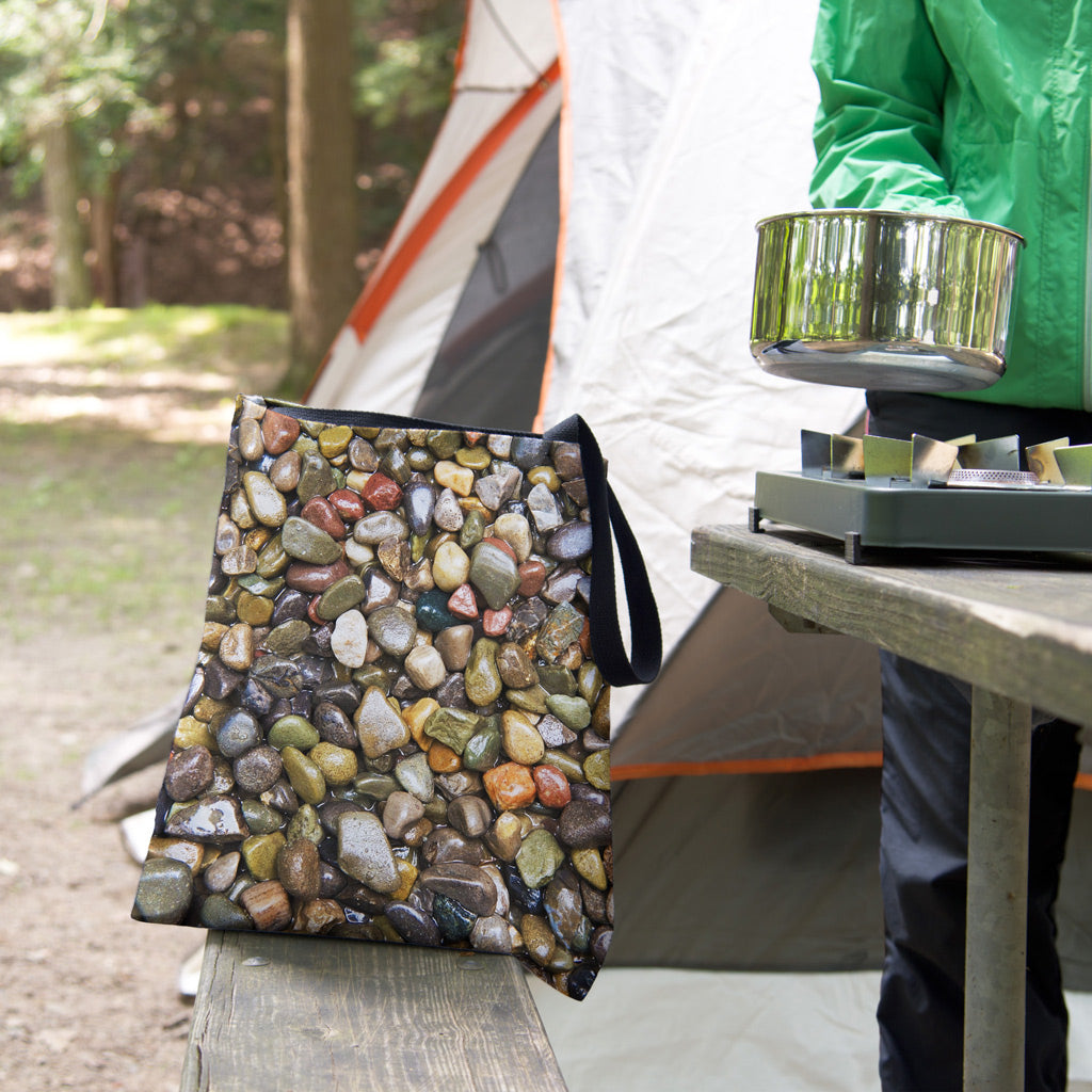 A tote bag featuring the pebbles and stones design sits on the bench of a campsite picnic table as the camper cooks.