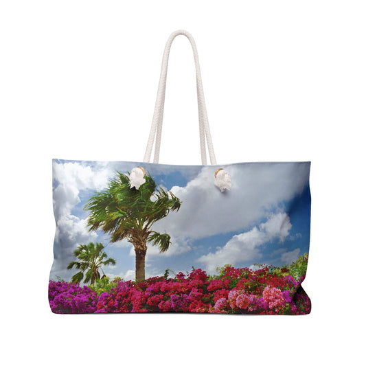 Front view of a beach bag featuring vibrant photo art of a bright blue sky, windy palm trees and brilliant tropical flowers.