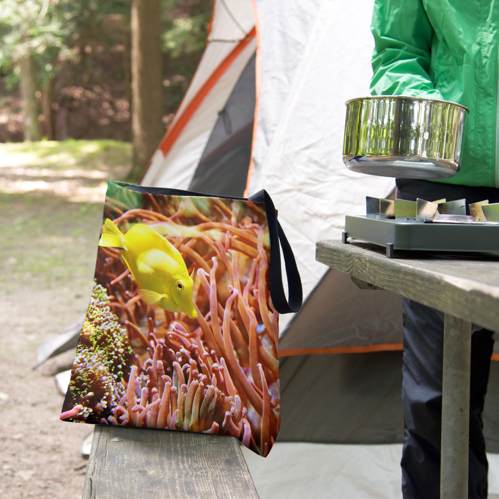 A tote bag featuring the yellow fish design sits on the bench of a campsite picnic table as the camper cooks.